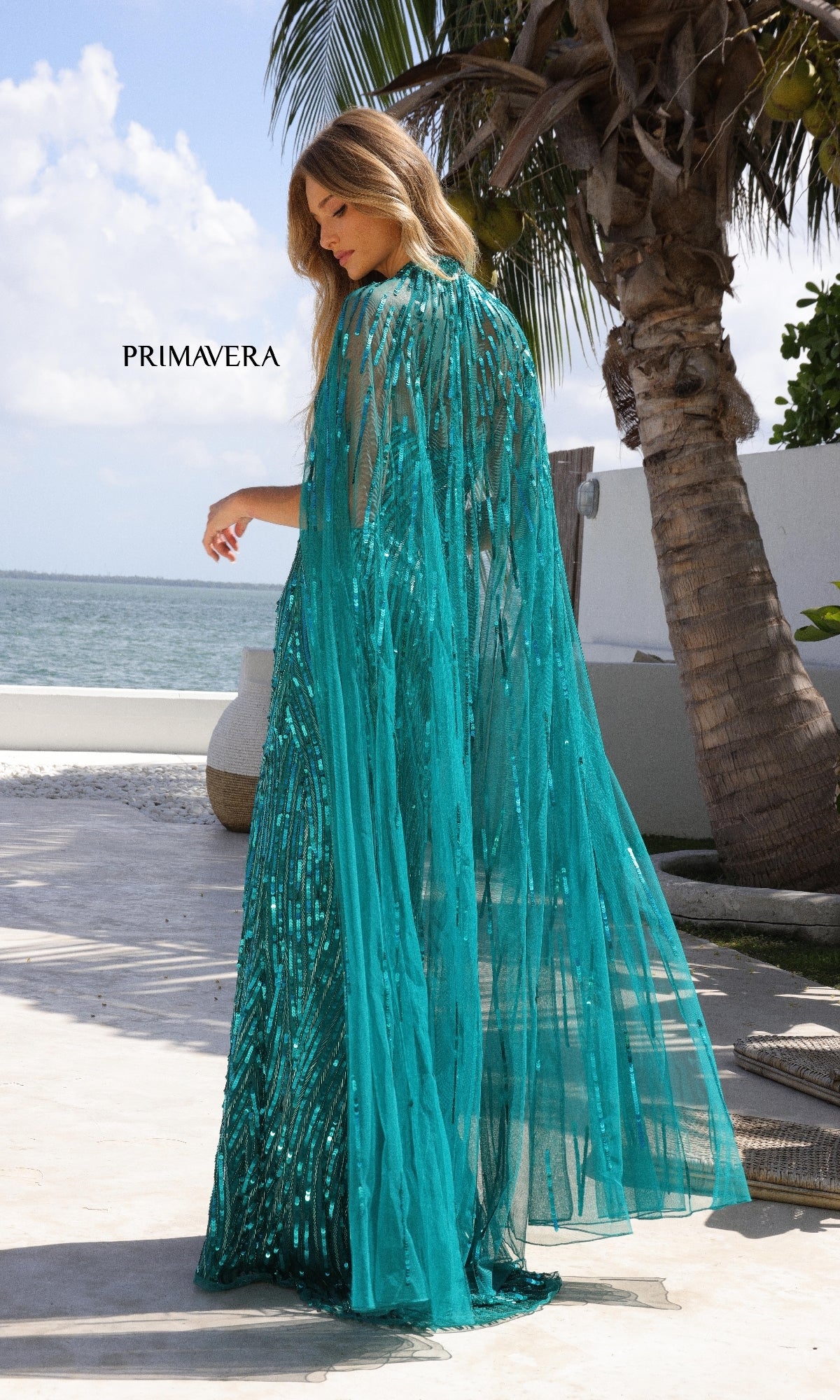 A woman stands outdoors near palm trees and water, facing away from the camera, wearing the Primavera 4376—a teal, sequined long prom dress with a sheer cape.
